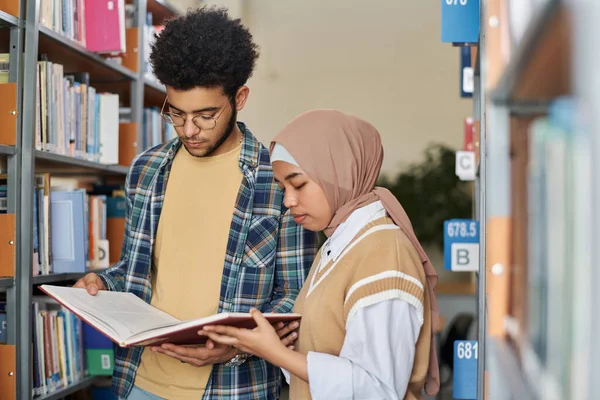 書棚の近くに立って図書館で一緒に本を読む多民族学生のカップル — ストック写真