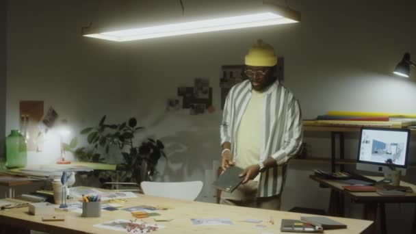 アフリカ系アメリカ人のデザイナーは夕方にオフィスを歩き 机に座ってコピーブックを描く — ストック動画