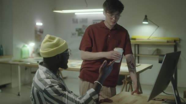 コーヒーカップを持っている若いアジアのデザイナーとアフリカ系アメリカ人の同僚とコンピュータ上でプロジェクトを議論しながら 一緒に遅くオフィスで働く — ストック動画