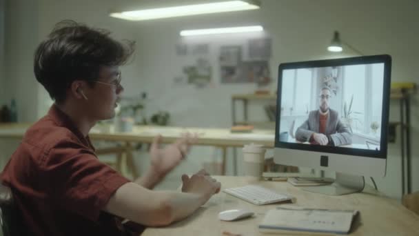 オフィスで仕事をしながら コンピュータ上のビデオ通話を介して男性の同僚と話す無線イヤフォンの若いアジアのデザイナー — ストック動画