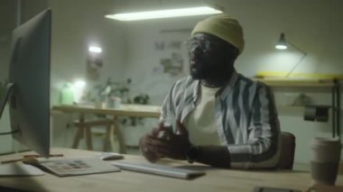 Afrika kökenli Amerikalı erkek tasarımcı ofiste geç saatlere kadar çalışırken bilgisayar üzerinden el sallıyor ve konuşuyor.