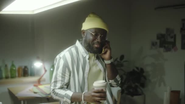 非洲裔美国人设计师高高地坐在桌子上 手握咖啡杯 晚上在工作室里用手机交谈的照片 — 图库视频影像