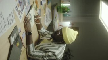 Afrikalı Amerikalı erkek tasarımcının gece boyunca yaratıcı stüdyoda çalışırken müzik dinleyip çizim yaptığı dikey bir fotoğraf.