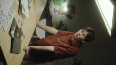 Genç Asyalı tasarımcının gece yaratıcı stüdyoda çalışırken üzerinde kağıt kolajlar, eskiz defteri ve renk paleti olan masasında poz verirken dikey çekimleri.