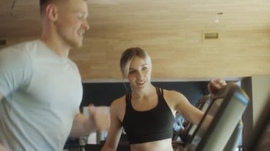 Spor yaparken koşu bandında koşan neşeli bayan antrenör adamla konuşuyor.