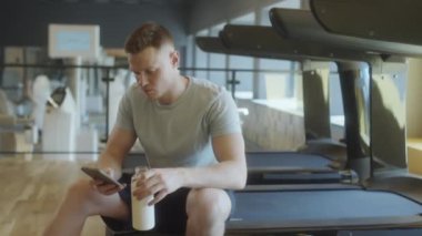 Spor giyim sektöründe kaslı bir adam spor salonunda koşu bandında oturmuş, elinde su şişesi ve akıllı telefondan mesajlar tutarken idmandan sonra dinleniyor.