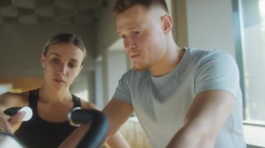 Spor salonunda bisiklete binen ve spor bisikletini nasıl kullanacağını anlatan kadın antrenörü dinleyen bir adam.