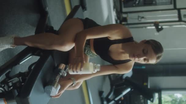 穿着运动服的妇女在运动后躺在健身房的长椅上 用瓶子喝水的垂直镜头 — 图库视频影像