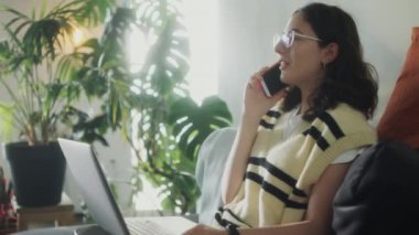 Z jenerasyonundaki bir kızın evde dizüstü bilgisayarıyla oturup uzaktan çalışırken gülümsediği ve telefonla konuştuğu orta boy bir fotoğraf.