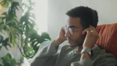 Genç yakışıklı adam evdeki kanepede oturuyor, kablosuz kulaklık takıyor ve başını müziğe çeviriyor.