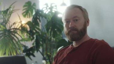 Evde dizüstü bilgisayarla otururken kameraya gülümseyerek poz veren sakallı adamın göğüs görüntüsü.