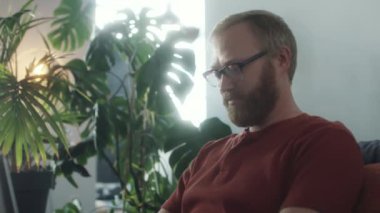 Genç sakallı adamın kanepede oturup, internet üzerinden konuşurken ve evde çalışırken bilgisayarında yazarken çekilen görüntüler.