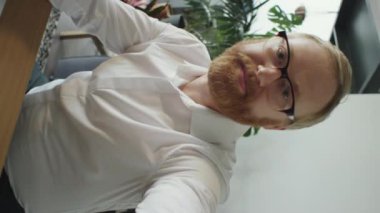 Beyaz sakallı beyaz iş adamının resmi gömlek ve gözlüklü görüntüsü çevrimiçi video görüşmesi üzerinden kameraya el sallıyor ve konuşuyor.