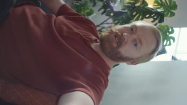 Kızıl sakallı adamın kolları açık bir şekilde akıllı telefonu tutarken ve evdeki video konuşmalarıyla konuşurken dikey görüntüsü.