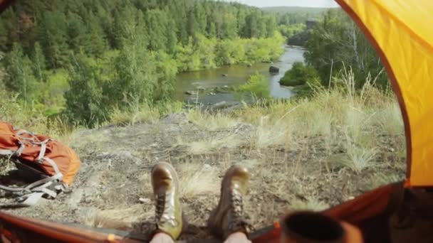 Povラックフォーカスショットの女性観光客でブーツに座ってテントでキャンプ場 保持ティーカップと見て風光明媚な川と緑の森 — ストック動画