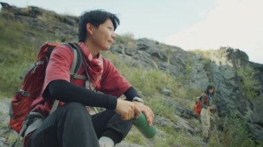 Asya 'lı kadın turistin yaz yürüyüşü sırasında nehir kıyısında bir şişe suyla otururken ve kız arkadaşıyla konuşurken PAN fotoğrafı.