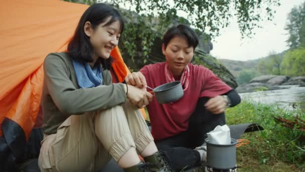两名亚洲女游客在河岸的营地吃晚餐时 把铝箔袋中的热食物放进锅里 — 图库视频影像