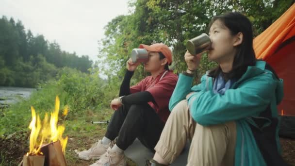 川沿いのキャンプファイヤーの近くに座っている2人のアジアの女性観光客 お茶を飲むと周りの美しい自然について話す — ストック動画