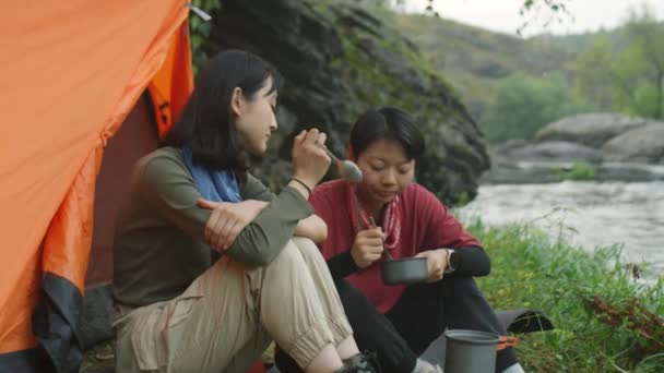 两名亚洲女游客坐在帐篷旁 边吃边聊天 边在河边露营 — 图库视频影像