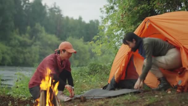 二人のアジアの女性観光客はそれを維持しながら たき火で木製の小枝を投げます川岸のキャンプ場で燃焼 — ストック動画