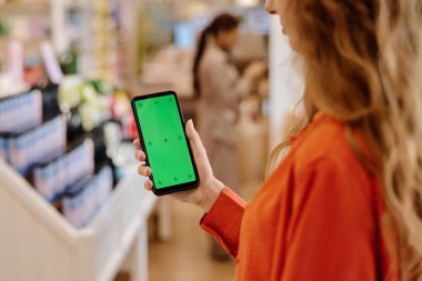 Alışveriş merkezinde alışveriş yaparken akıllı telefonunda indirimli uygulama kullanan genç bir kadın.