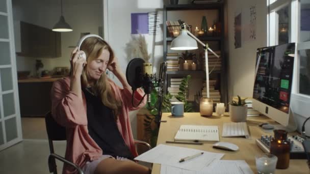 ズームで撮影した女性ブロガーを置くヘッドフォンとマイクで話している間に録音ポッドキャストで居心地の良いホームスタジオでキャンドルで飾られた — ストック動画