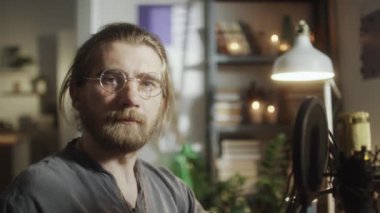 Evdeki kayıt stüdyosunda mikrofonun yanında oturmuş kameraya poz veren sakallı genç adamın portresi.