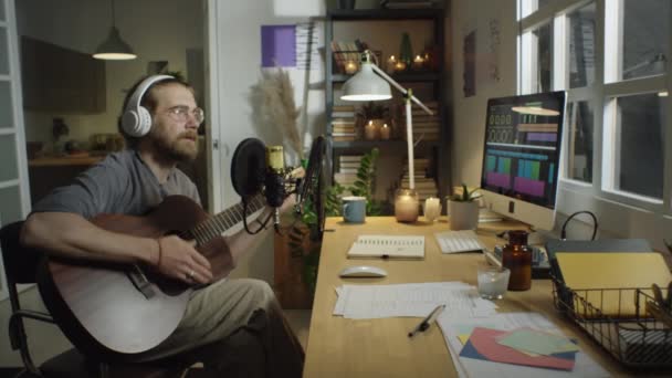 ヘッドフォンで男性音楽家のショットでズームギターを演奏し 自宅のレコーディングスタジオでマイクで歌う — ストック動画