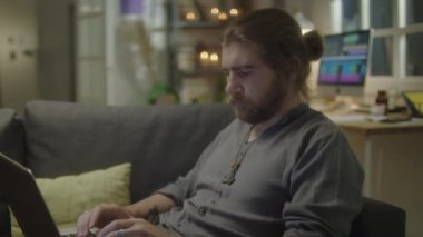 Genç sakallı adamın kanepede oturup dizüstü bilgisayarda yazarken, internette gezinirken ya da akşam evde uzaktan çalışırken görüntüsünü kaldır.