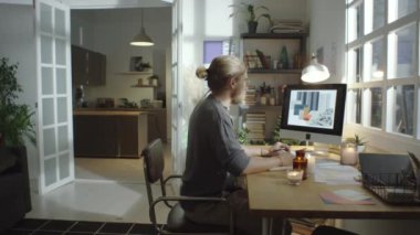 Sakallı erkek tasarımcının bilgisayarda iç planları izlerken ve evde rahat bir atmosferde çalışırken elinde mumlarla masa başında notlar alırken görüntüsünü yakınlaştır.
