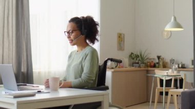 Tekerlekli sandalyede oturan genç iş kadını, evden çalışırken internetten bilgisayarla el sallıyor ve konuşuyor.