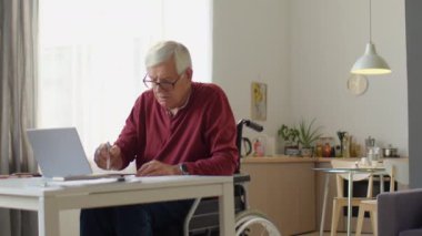 Üst düzey işadamının tekerlekli sandalyede oturup dizüstü bilgisayarda yazarken ve evden uzaktan çalışırken notlar alırken orta uzunlukta bir fotoğrafı.
