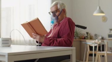 Tekerlekli sandalyede oturan, nebulizör kullanan ve evde kitap okuyan yaşlı bir adam.