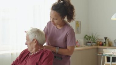 Kadın fizyoterapist, tekerlekli sandalyedeki yaşlı adamın başını döndürüyor ve ona evde tıbbi bakım sağlarken konuşuyor.