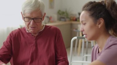 Gözlüklü yaşlı adamın dizüstü bilgisayarla oturduğu ve evdeki kadın bakıcı ile belgeleri tartıştığı el kamerasıyla çekilmiş bir fotoğraf.