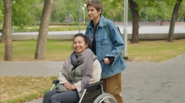 Orta uzunlukta, neşeli genç bir kadının, kadın arkadaşını tekerlekli sandalyeye iterken ve onunla sonbahar günü parkta yürürken sohbet ederken görüntüsü.