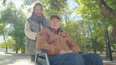 Young woman pushing senior grandfather in wheelchair and telling something while walking in park on sunny day