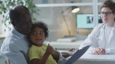 African American dad sitting with little son in clinic, reading medical agreement and talking with pediatrician