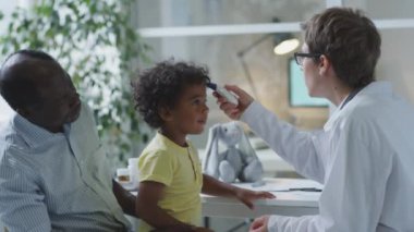 Küçük Afrikalı Amerikalı çocuk babasıyla otururken kadın çocuk doktoru klinikte kontrol sırasında ateşini kızılötesi termometreyle ölçüyor.