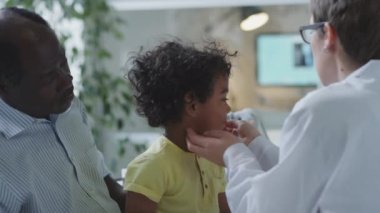 Küçük Afrikalı Amerikalı çocuk babasıyla otururken, kadın doktor pediatri kliniğinde sağlık kontrolü sırasında ensesinde lenf düğümlerini inceliyor.