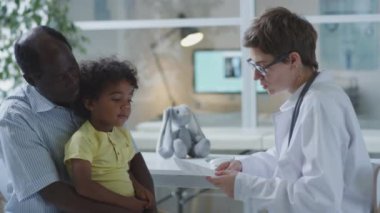 Reçeteli ilaç tutan ve tedavi planını Afrikalı Amerikalı babaya açıklayan kadın çocuk doktoru küçük oğluyla klinikte danışmanlık yapıyor.