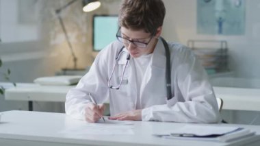 Beyaz önlüklü ve gözlüklü genç bir kadın doktorun portresi masaya notlar yazıyor ve sonra da klinikte çalışırken kameraya poz veriyor.