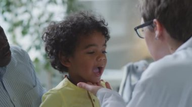 Küçük Afrikalı Amerikalı çocuk babasıyla otururken kadın doktor da pediatri kliniğinde kontrol sırasında boğazını fenerle kontrol ediyor ve lenf düğümlerini inceliyor.