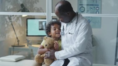 Sevimli Afro-Amerikan çocuk elinde ayı oyuncağı tutarken ve klinikteki kanepede birlikte otururken dikkatli bir çocuk doktoruyla konuşurken.