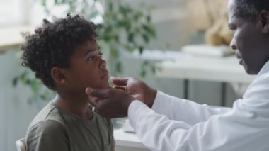 Küçük Afrikalı Amerikalı çocuk pediatri kliniğinde otururken erkek doktor kontrol sırasında fenerle boğazını muayene ediyor ve lenf bezlerine dokunuyor.