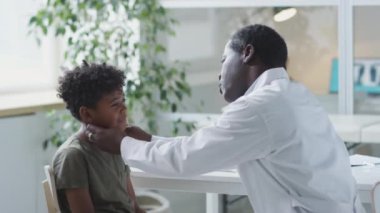 Afrikalı Amerikalı çocuk doktoru küçük çocuğun lenf bezlerini inceliyor ve klinikte fenerle boğazını kontrol ediyor.