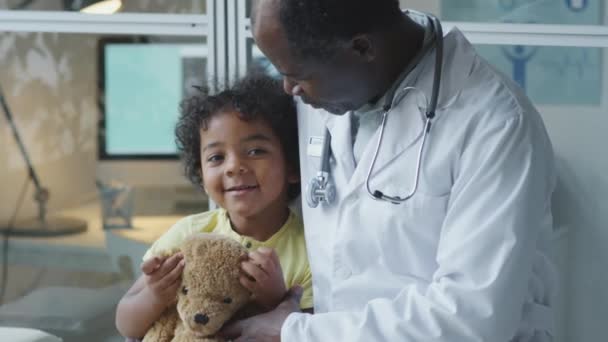 アフリカ系アメリカ人の小児科医は 熊のおもちゃで座ってクリニックで彼と話す小さな子供を受け入れる — ストック動画