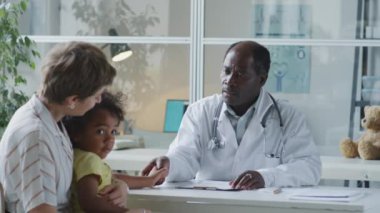 African American pediatrician holding hand of little child and speaking with his mother during consultation in clinic