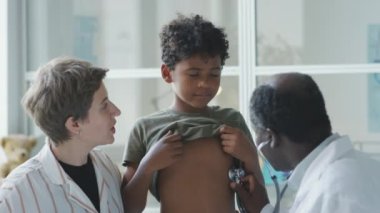 Pediyatrist onu steteskopla muayene ederken ve klinikte kontrol sırasında annesine sarılırken Afro-Amerikan çocuk tişörtünü kaldırdı.
