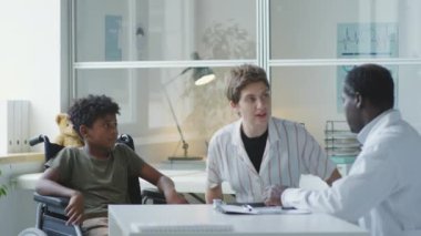 Anne ve küçük çocuk tekerlekli sandalyede kliniği ziyaret ediyor ve Afrikalı Amerikalı çocuk doktoruna danışıyorlar.